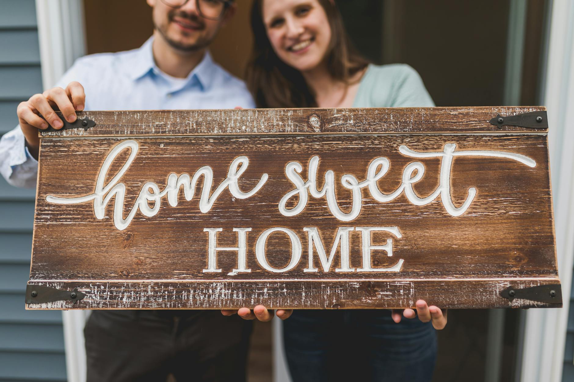 Couple smiling, holding a 'Home Sweet Home' sign, standing in a cheerful outdoor setting.