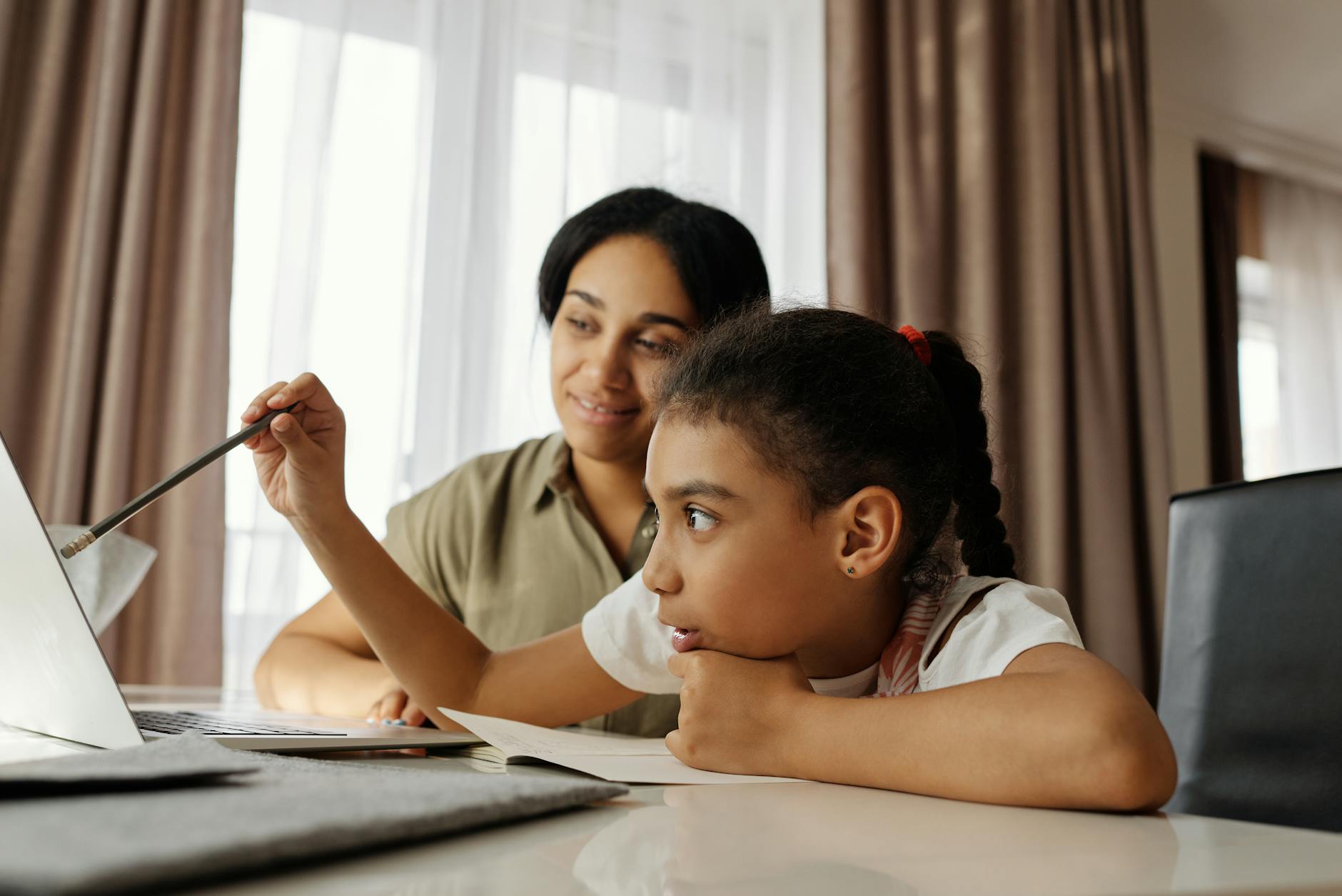 Mother and daughter learning together at home, focused on a laptop, surrounded by books and a cozy atmosphere.