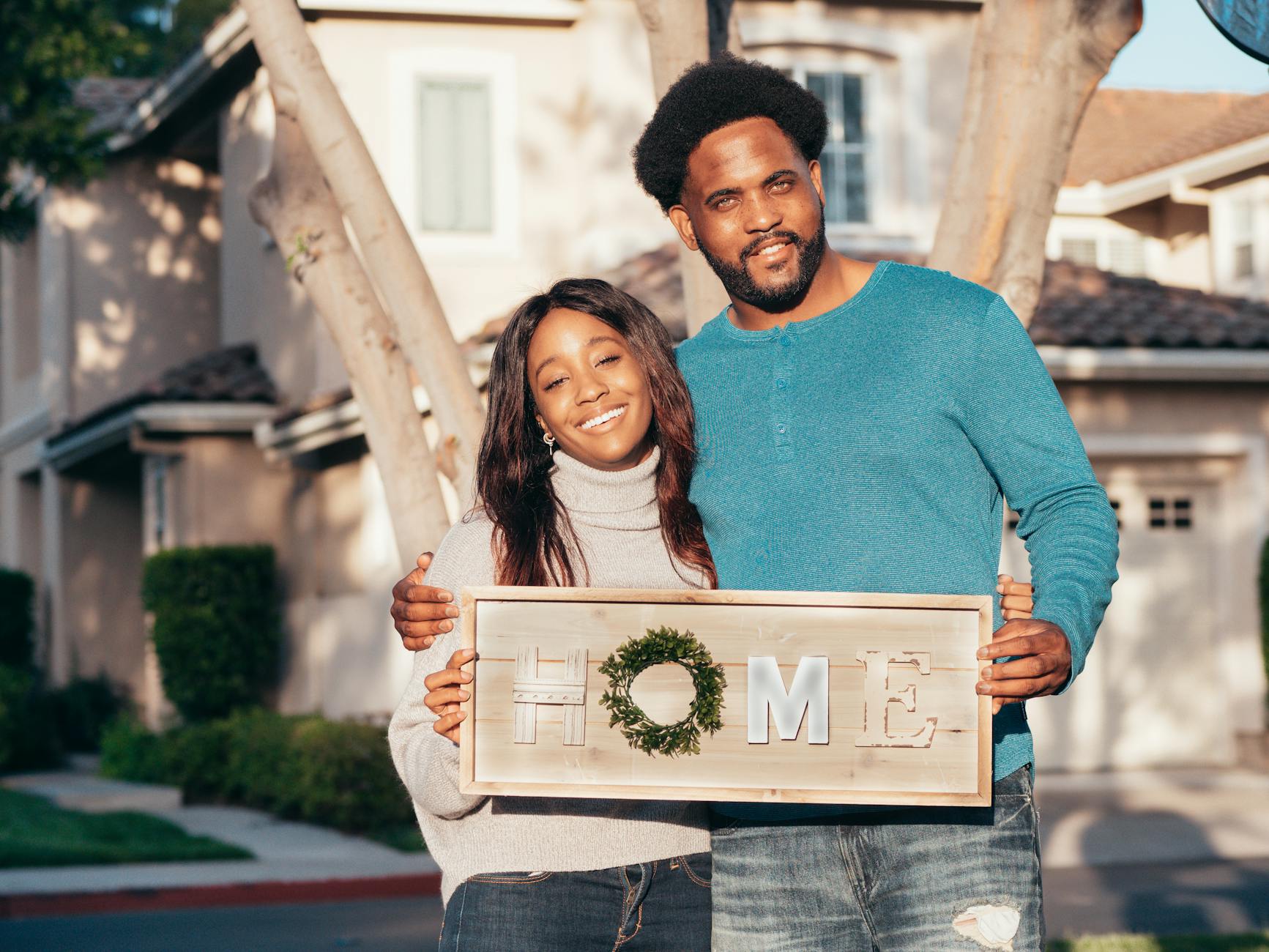 Happy couple smiling and holding a 'Home' sign in front of their new house.