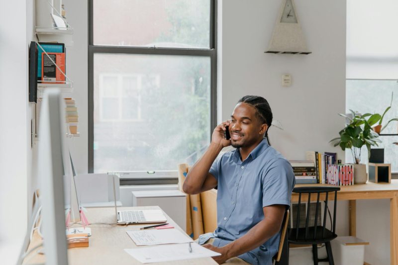 Adult man in a professional office setting, talking on a phone while working at a desk.