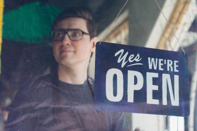 Smiling adult man stands behind glass with a welcoming "Yes, We're Open" sign in a store setting.