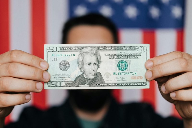 Adult man obscuring his face with a dollar bill against a blurred background of the American flag.