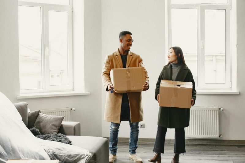 Smiling couple carrying cardboard boxes into a bright, empty home, ready to move in.