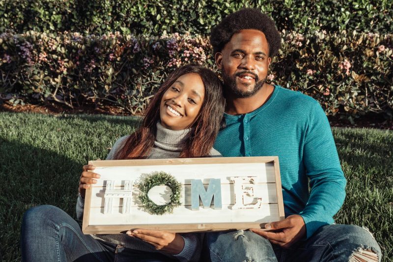 Smiling couple sitting on a lawn, holding a colorful 'Home' sign, celebrating their new home ownership.