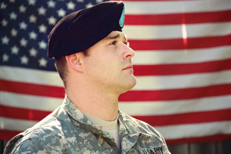 US soldier in uniform stands proudly with the American flag waving in the background.