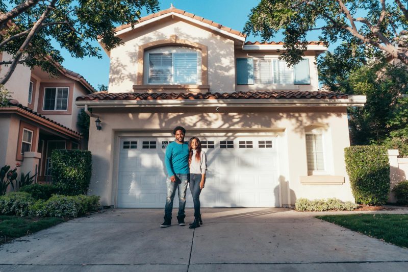 Smiling couple stands outside their newly purchased house on a sunny day.