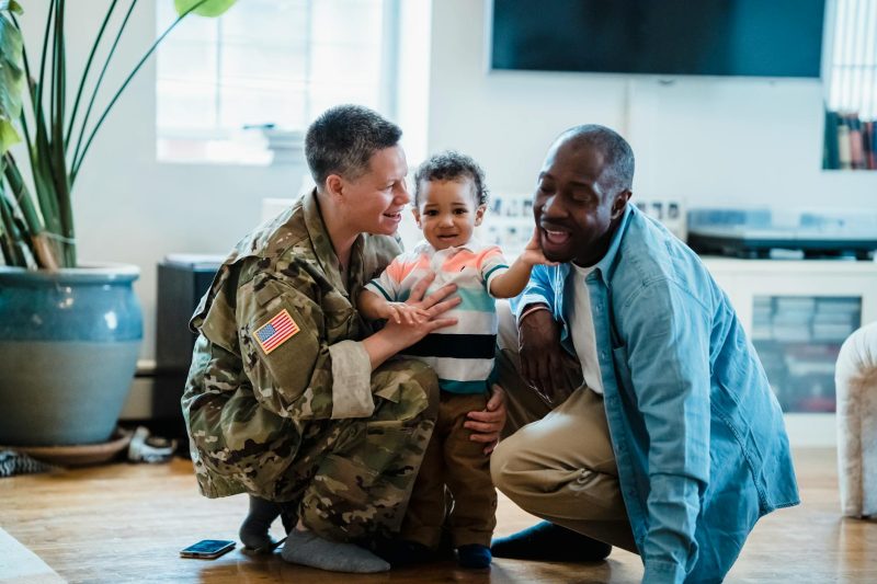 A military parent embraces their partner and child indoors, sharing a joyful family moment.